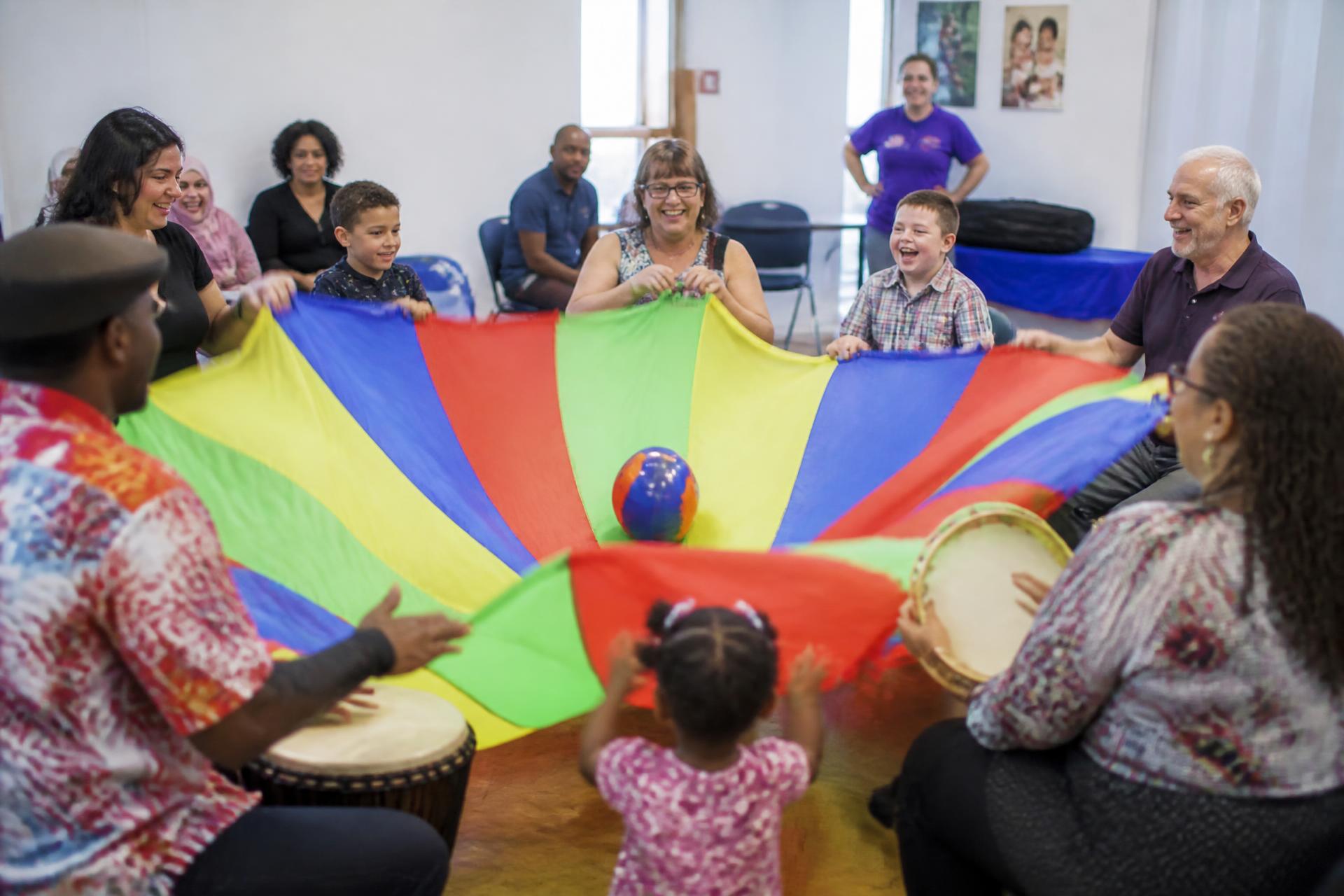 Grupo diverso de personas participando en una actividad recreativa con paracaídas de colores e instrumentos musicales